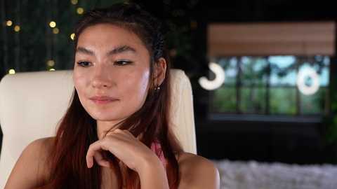 Media: Video of an East Asian woman with long, straight brown hair styled in two braids, wearing a pink top, sitting on a white chair indoors, focused on a pink object in her hand, blurred background with greenery and a window.