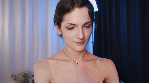 Media: Video of a young woman with fair skin, dark hair, and delicate features, wearing a thin-strapped, light-colored dress. Background features soft lighting and dark curtains.