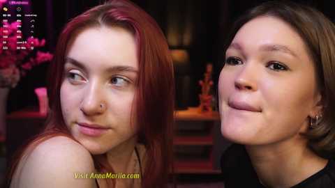 Media: Video of two young women with pale skin and light brown hair, one with a nose ring, smiling and sitting closely. Background shows dimly lit room with blurred objects.