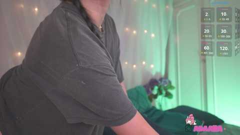 Media: A video of a curvy, plus-sized woman with medium brown skin and long, dark hair, wearing a dark gray t-shirt, standing in a cozy bedroom with fairy lights, a blue bed, and a pink logo in the corner.