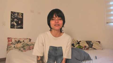 Media: Video of a young person with short dark hair, wearing a white t-shirt with a graphic and denim overalls, sitting on a bed with floral pillows and a poster of a band on a beige wall.