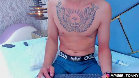 Media: Video of a shirtless man with tattoos, including a large angel wings design on his chest, wearing blue Adidas pants. He's sitting on a bed with white sheets, blue pillow, and a laptop.