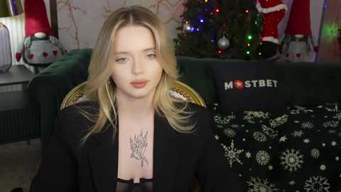 Media: Video of a young, fair-skinned, blonde woman in a black blazer with a tattoo on her chest, sitting in a cozy, festive living room with a decorated Christmas tree and holiday decor.