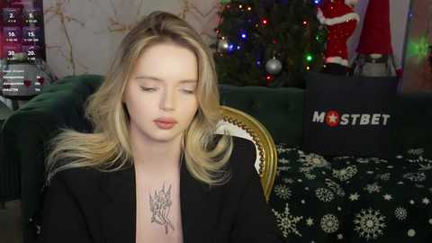 Media: Video of a blonde woman with a black tattoo on her chest, wearing a black top, sitting in a green velvet armchair, surrounded by a decorated Christmas tree and a \"Mostbet\" banner in the background.