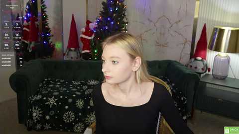 Media: Video of a young, fair-skinned blonde woman in a black top, sitting indoors near a Christmas tree, decorated with red and white ornaments, and a green couch with a festive blanket.