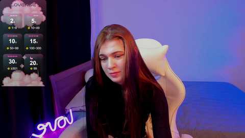 Media: A video of a young, fair-skinned woman with long, straight brown hair, wearing a black top, sitting on a white cushioned chair in a dimly lit room with a blue and purple light glow.