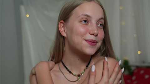 Video of a young, fair-skinned blonde woman with straight hair, wearing a black necklace, smiling, with soft-focus background featuring white curtains and festive lights.