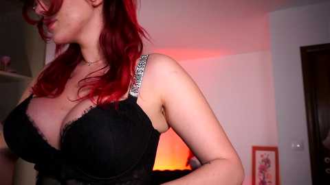 Video of a fair-skinned woman with red hair, wearing a black lace bra, in a dimly lit room with warm orange lighting.