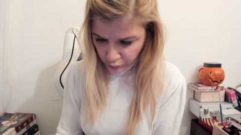 A video of a blonde woman in a white sweater, focused on a pumpkin-shaped container, surrounded by books and a cluttered desk.