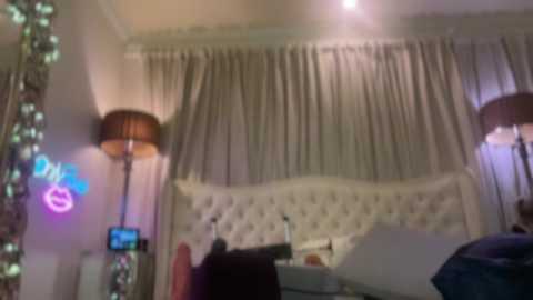 Video of a dimly lit, modern bedroom with a tufted white headboard, beige curtains, and a large lamp.