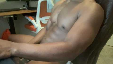 Media: Video of a shirtless, muscular man with a light skin tone sitting in an orange office chair, displaying defined abs and arms. Background includes a desk with a computer monitor and keyboard.