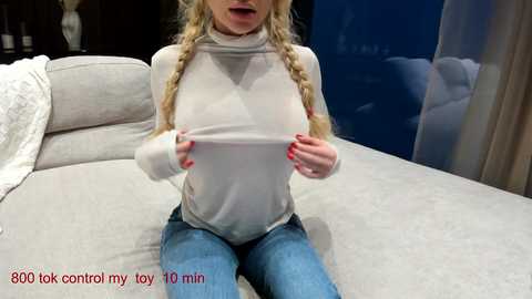 Video of a blonde woman with twin braids, wearing a sheer turtleneck, lifting her breasts, seated on a beige couch.