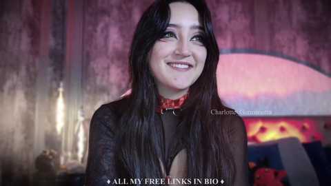 Media: Video of a smiling, fair-skinned woman with long black hair, wearing a sheer black top and a red choker, in a dimly lit room with a pink and purple sunset backdrop.