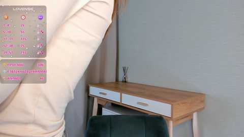 Media: Video of a person in beige pants, standing near a light wooden desk with two drawers, and a dark green chair, set against a gray wall with a small plant on top.