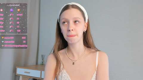 Media: Video of a young, slender Caucasian woman with long brown hair, wearing a white headband, delicate necklace, and a lace camisole, standing in a modern room with a calendar and desk.