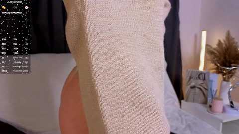 A video of a woman's torso in a beige, textured sweater, standing in a softly lit bedroom with a bed, potted plants, and a nightstand.