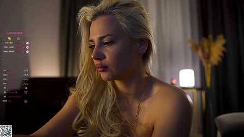 Video of a blonde woman with fair skin and long wavy hair, topless, seated indoors at night, with a dimly lit background featuring curtains and a table with a lamp.