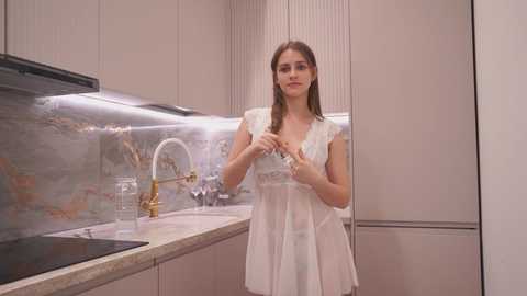 Media: Video of a young woman with long brown hair, wearing a sheer white dress, standing in a modern kitchen with beige cabinets, a stainless steel sink, and a black stove.