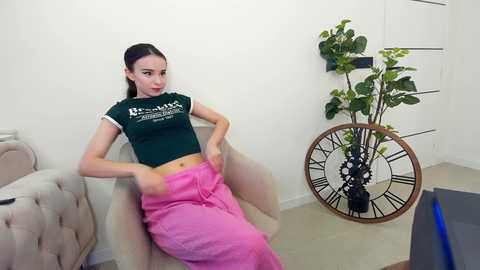 Media: Video of a slim, light-skinned woman with long brown hair, wearing a dark green crop top and bright pink pants, sitting on a beige armchair. A decorative clock with green leaves and a metal stand is on the right.