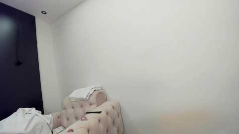 Media: Video of a minimalist bedroom with a tufted beige headboard, a white bed with messy sheets, and a dark wall with a recessed light.