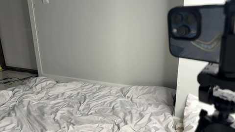 Media: A video of a messy, unmade bed in a dimly lit room, with a smartphone capturing the scene, partially reflecting the room's white walls and a partially visible person in the background.