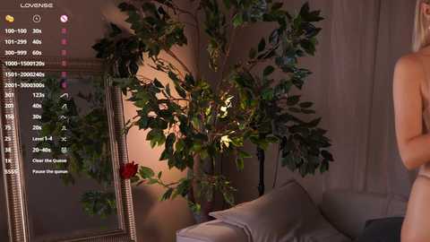 Video of a nude woman with fair skin standing beside a large fake plant in a dimly lit living room.