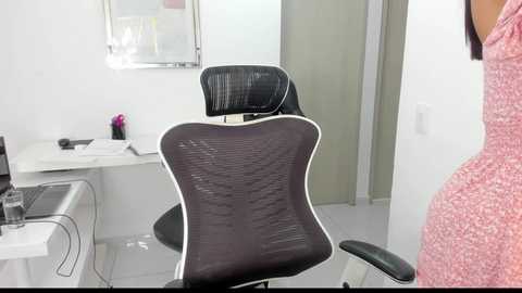 Media: Video of a modern office chair with a black mesh backrest and armrests, placed in a minimalist, white-walled room with a desk, computer monitor, and a pink sweater visible on the right.