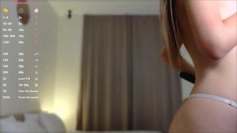 Media: A video of a topless woman with light skin, wearing a grey thong, standing in a dimly lit room with beige curtains. A digital interface with camera settings is overlaid on the left side.