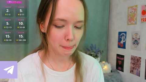 Media: Video of a young, fair-skinned woman with brown hair, wearing a white shirt, in a cozy room with pastel-colored walls and framed art, with a digital clock display in the top left.