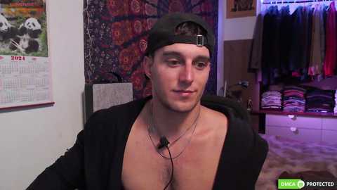 Media: Video of a shirtless, light-skinned young man with a fit physique, wearing a black hoodie and backward cap, with a necklace and earphones. Background features a colorful tapestry, a calendar, and a closet filled with clothes.