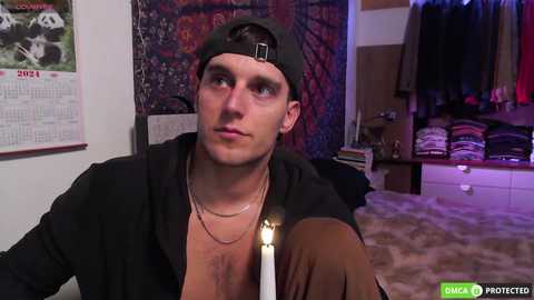 Media: Video of a young Caucasian man with short brown hair, wearing a black baseball cap, black jacket, and multiple gold chains, seated in a dimly lit room with a calendar, hanging tapestry, and clothing racks in the background.