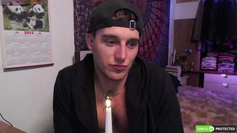 Media: Video of a young, fair-skinned man with a slim build wearing a backward black baseball cap and a black hoodie, holding a lit candle in a cozy room with a calendar, tapestry, and shelves in the background.