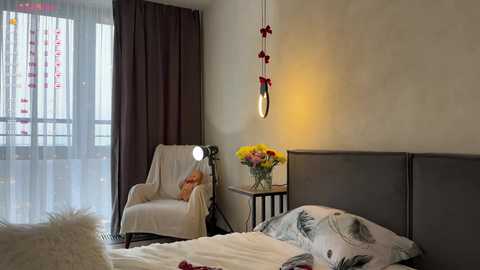 Media: Video of a minimalist, modern bedroom with a grey upholstered headboard, white bed, white chair, and a small nightstand holding a vase of flowers. A dangling light fixture with red accents hangs above the bed, casting a warm glow.