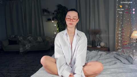 Media: A video of a slender, fair-skinned woman with glasses, sitting cross-legged on a bed, wearing a loose white shirt, in a dimly lit, modern living room with plants and a lamp.