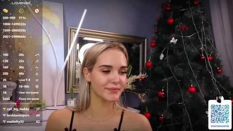 Media: Video of a blonde woman with fair skin, wearing a black top, smiling in a room with a Christmas tree decorated with red ornaments and lights, a framed picture, and a window.