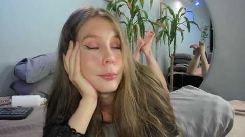 Media: Video of a fair-skinned, long-haired young woman with closed eyes, resting on a bed with a gray blanket. Behind her, a potted plant and a mirror reflect her legs, suggesting a cozy, minimalist room.