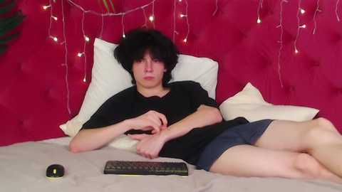 Video of a young person with short, black hair, lying on a bed with a red, tufted headboard, wearing a black shirt and shorts, holding a computer mouse on beige bedding.