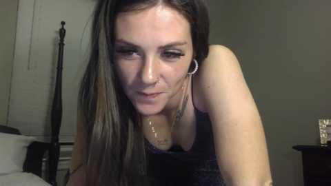 Media: Video of a fair-skinned woman with long brown hair, wearing a black tank top and silver hoop earrings, leaning forward with a serious expression. The background includes a white door, a black bed frame, and a nightstand.