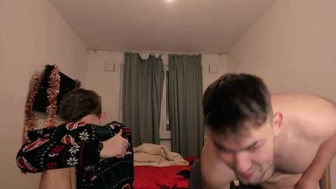 Media: A video of a shirtless man with short dark hair and a woman with brown hair wearing a festive sweater, standing in a dimly lit bedroom with beige walls, a bed, and curtains.