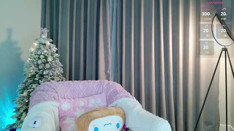 A video of a cozy, holiday-themed room with a snow-covered Christmas tree, plush pink armchair adorned with a white, fluffy blanket, and a large, modern tripod lamp.