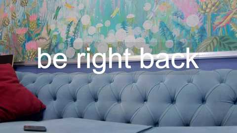 Media: A video of a cozy room with a blue, tufted velvet sofa, a vibrant, floral-patterned wallpaper, and a red pillow. Text reads \"be right back\" in white.