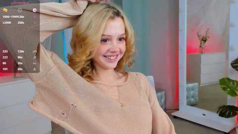 Media: Video of a blonde woman with fair skin, smiling, lifting a beige sweater. Background includes modern furniture, plants, and soft lighting.