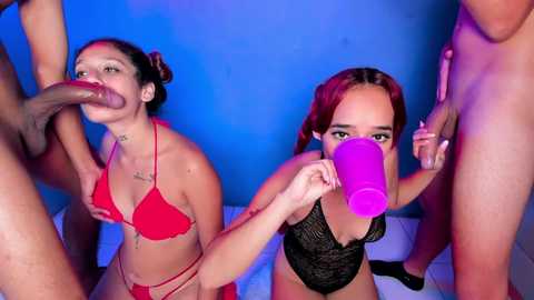 Media: Video: Two young women, one in a red bikini, the other in black lace lingerie, perform oral sex on two nude men; vivid blue background.