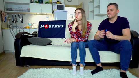 Media: Video of a young couple watching TV in a modern, minimalist kitchen with a white fridge and wooden floor. The woman is blonde, in a red plaid shirt, and the man is in a black shirt, both sitting on a cream sofa.