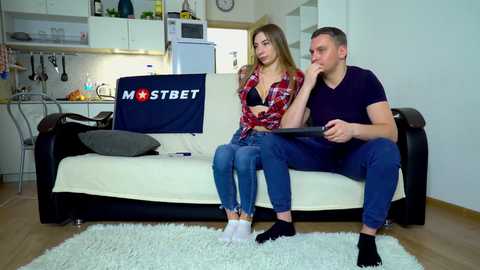 Video of a young, blonde woman in a red plaid shirt and jeans sitting on a sofa, looking thoughtful. Beside her, a young man in a dark shirt and jeans holds a tablet, also looking contemplative. Background shows a modern kitchen with white cabinets and stainless steel appliances.