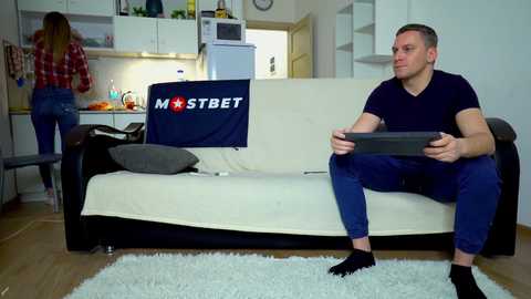 Media: Video of a man in a navy T-shirt and blue jeans sitting on a beige couch, holding a controller, while a woman in a red plaid shirt and jeans stands in a modern kitchen.