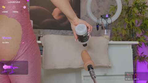 Media: Video of a woman in a pink, form-fitting dress, applying lotion to her lower back with a tan-colored dildo vibrator, set in a modern bathroom with a ring light, plants, and a bathtub.