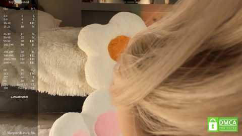 Media: Video of a blonde woman with shoulder-length hair, lying on a bed with a large, fluffy white dog. The bed is adorned with a large, white flower-shaped pillow.