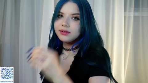 Media: Video of a young woman with long, teal hair, wearing a black choker and top, holding a phone, indoors with white curtains.