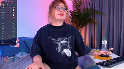 Media: Video of a plus-size woman with glasses and light skin, wearing a black T-shirt with a dog graphic, seated at a cluttered desk with a computer screen and water bottle.